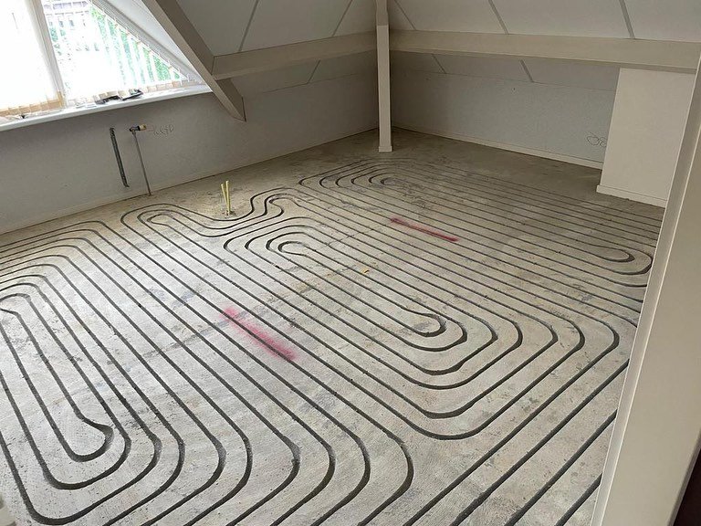 Vloerverwarming met slangenpatroon op de vloer in een lege ruimte, met leidingen en isolatiepanelen.
